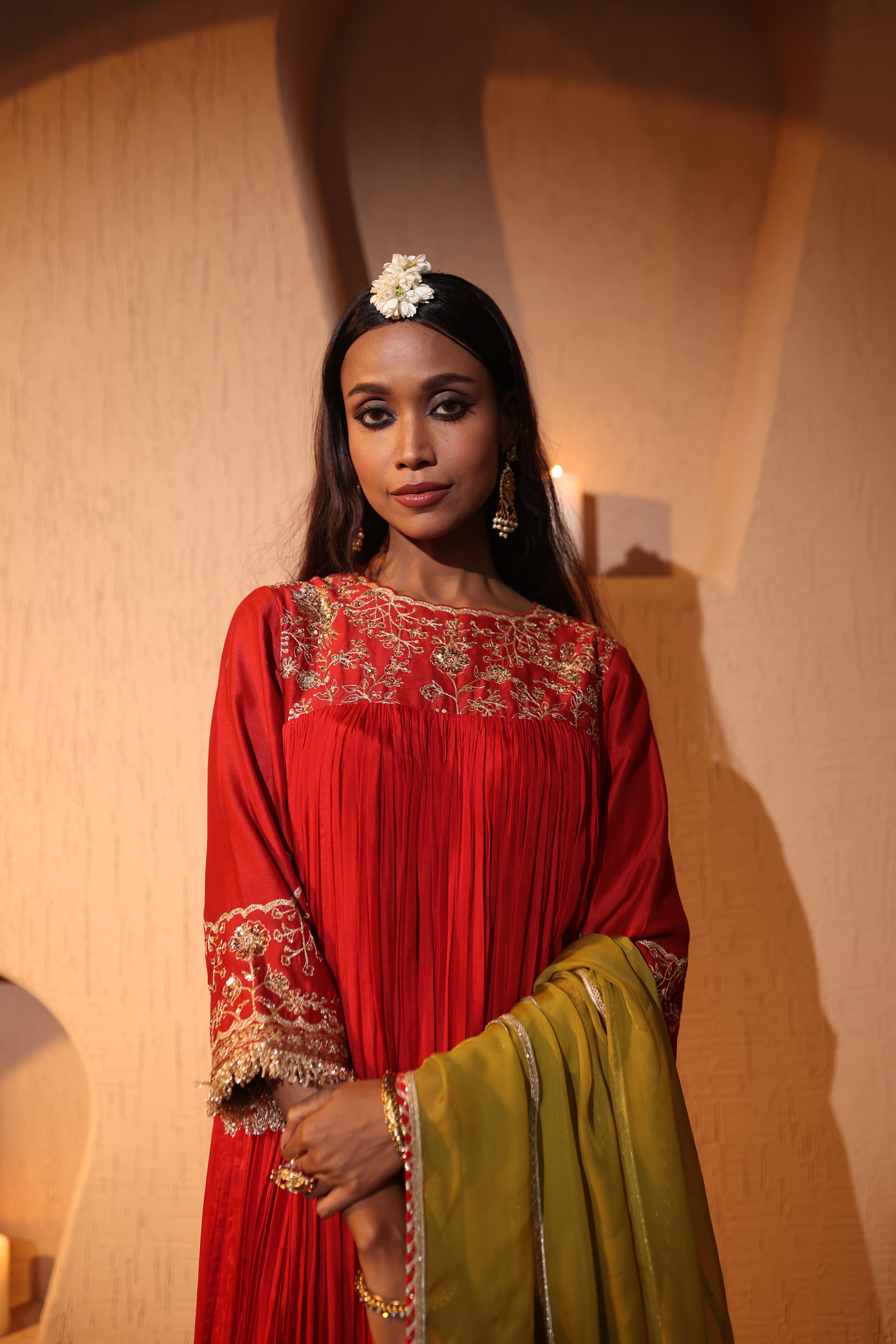 Woman in a red and green traditional outfit with floral headpiece against a beige wall.