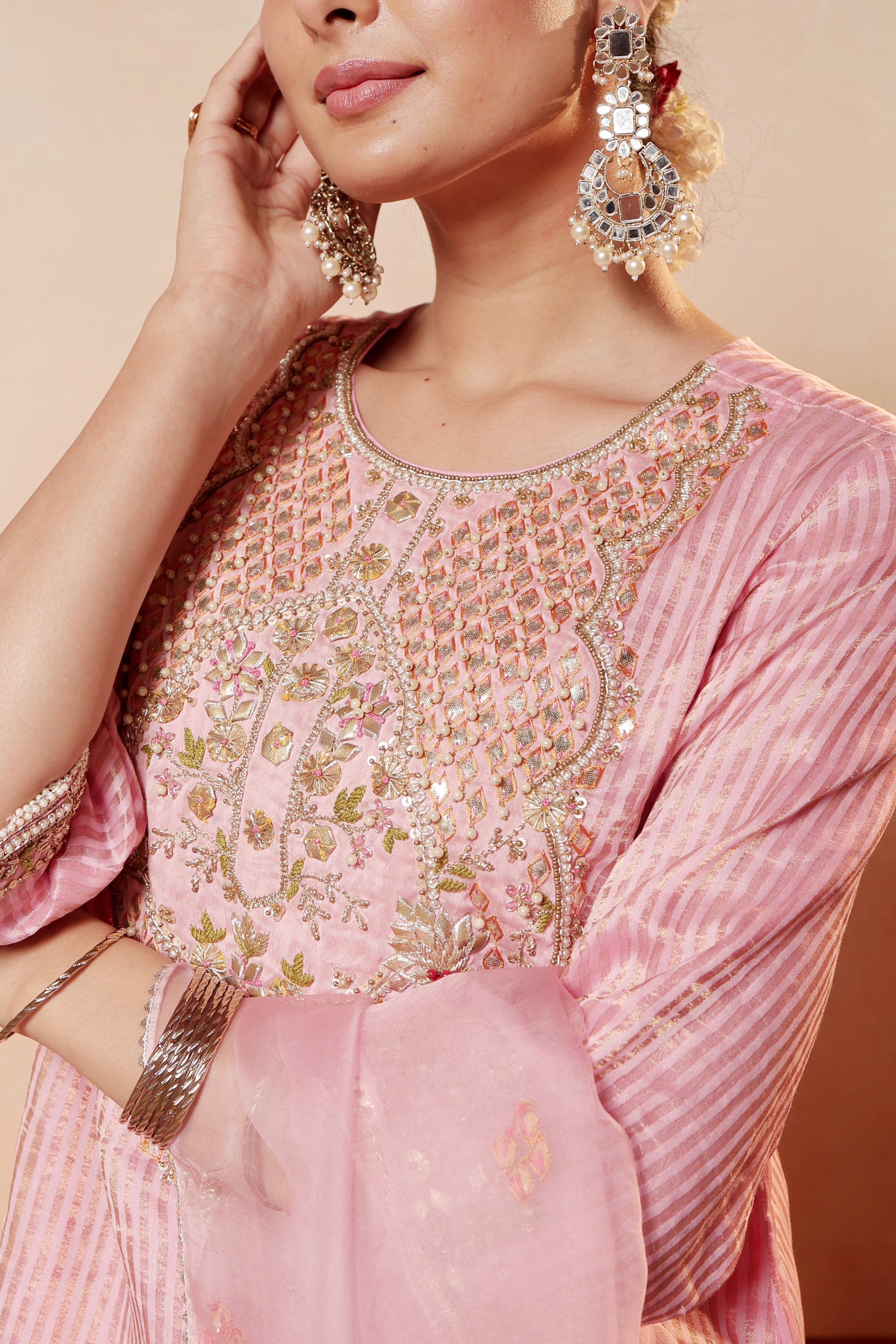 Close-up of a person wearing a pink embroidered outfit with jewelry on a beige background. Model wearing pink tissue silk kurta with intricate gota embroidery, paired with regal farshi salwar and dupatta – Sunehri Gota Collection by House of AAS.