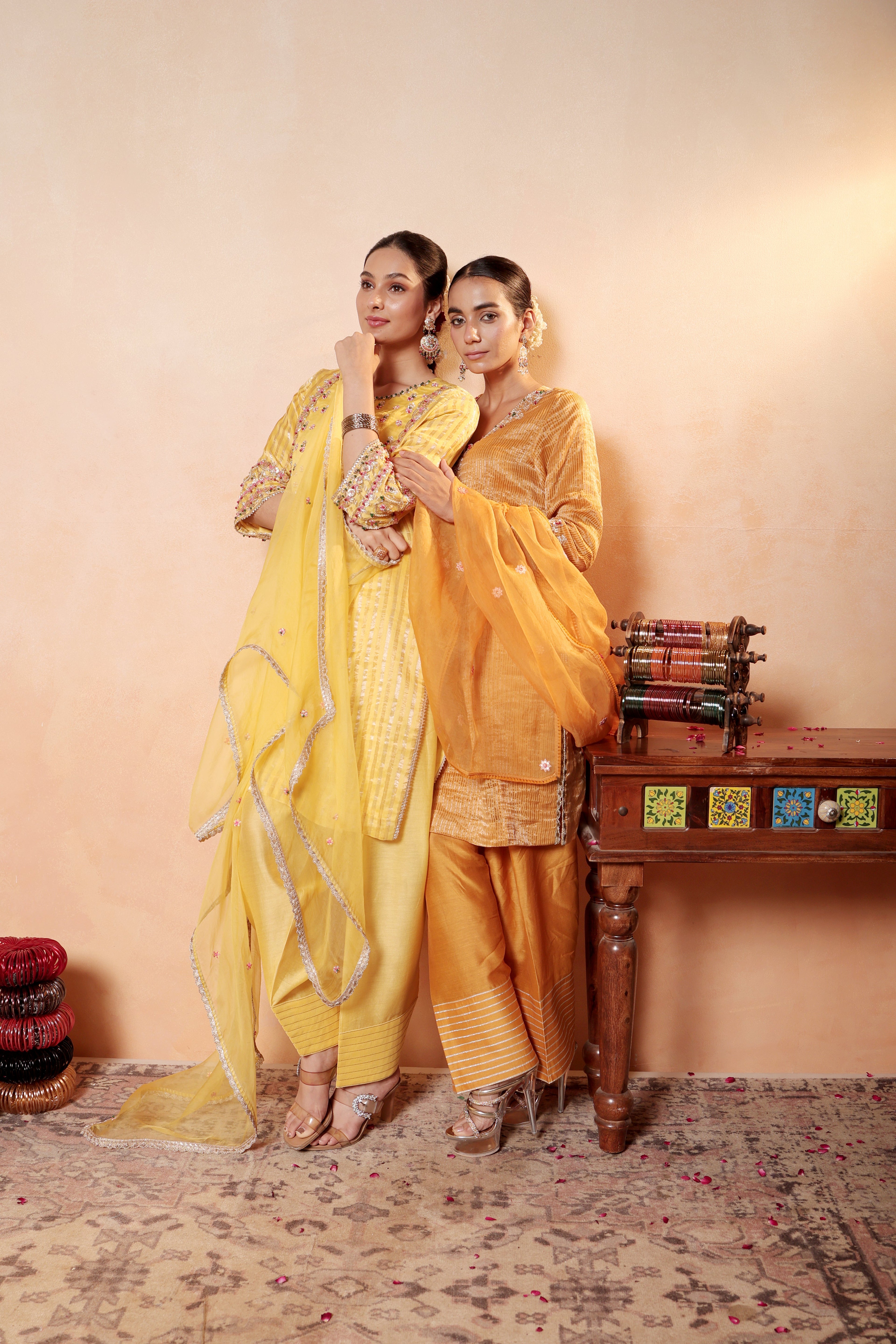 Two women in traditional yellow outfits standing in a room with a wooden table and decorative items. Model wearing orange silk striper tissue kurta with gota embroidery, paired with regal farshi salwar and dupatta – Sunehri Gota Collection by House of AAS.