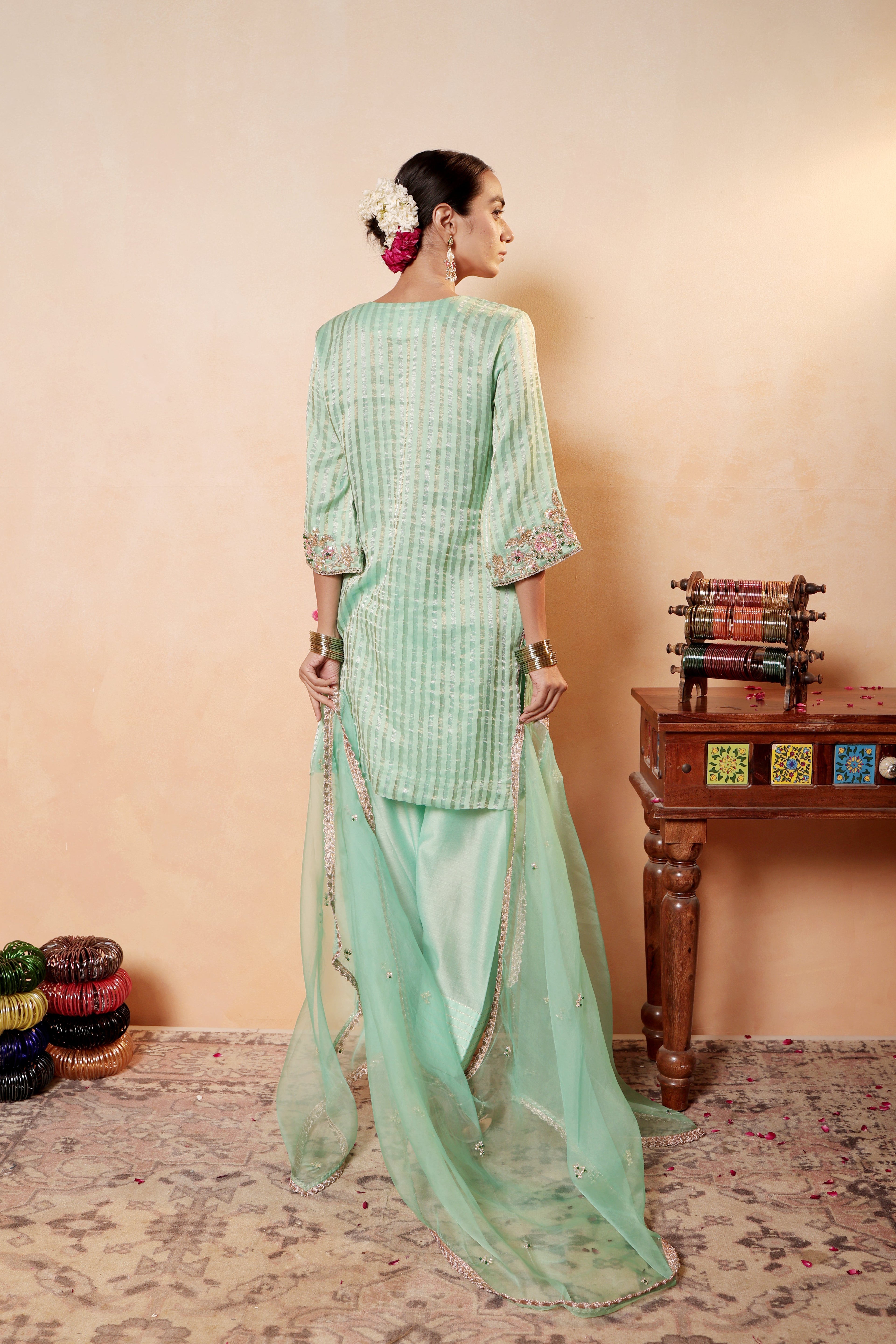 Woman in a light green traditional outfit standing against a beige wall with a wooden table and decorative items. Model wearing mint green silk striper tissue kurta with intricate gota embroidery, paired with regal farshi salwar and dupatta – Sunehri Gota Collection by House of AAS.