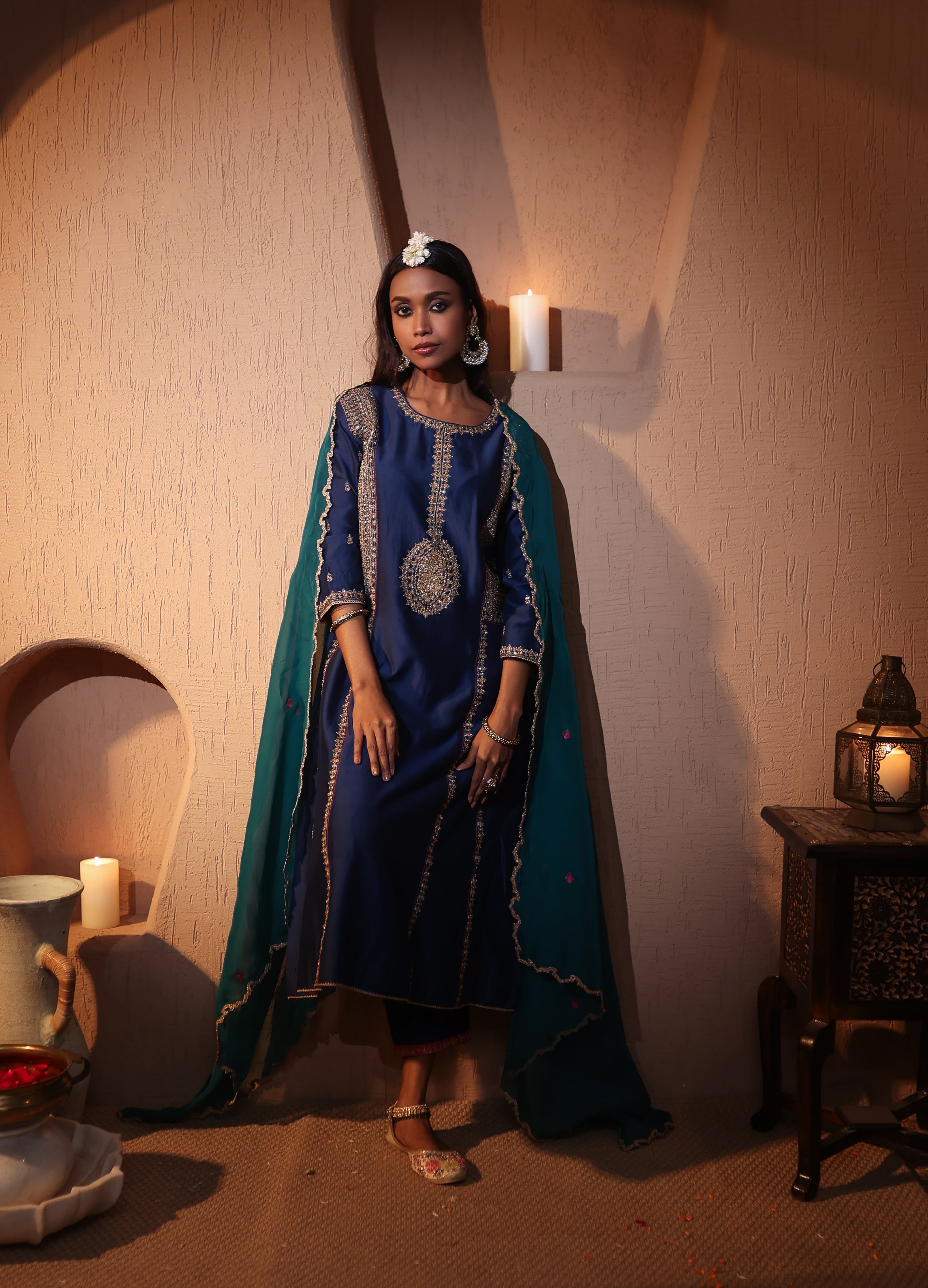 Woman in a blue silk chanderi skurta set with a emerald green organza dupatta standing in a warm-toned room with candles and decorative items.