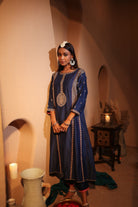 Woman in a blue traditional dress standing in a warm-toned room with a table and lamp.