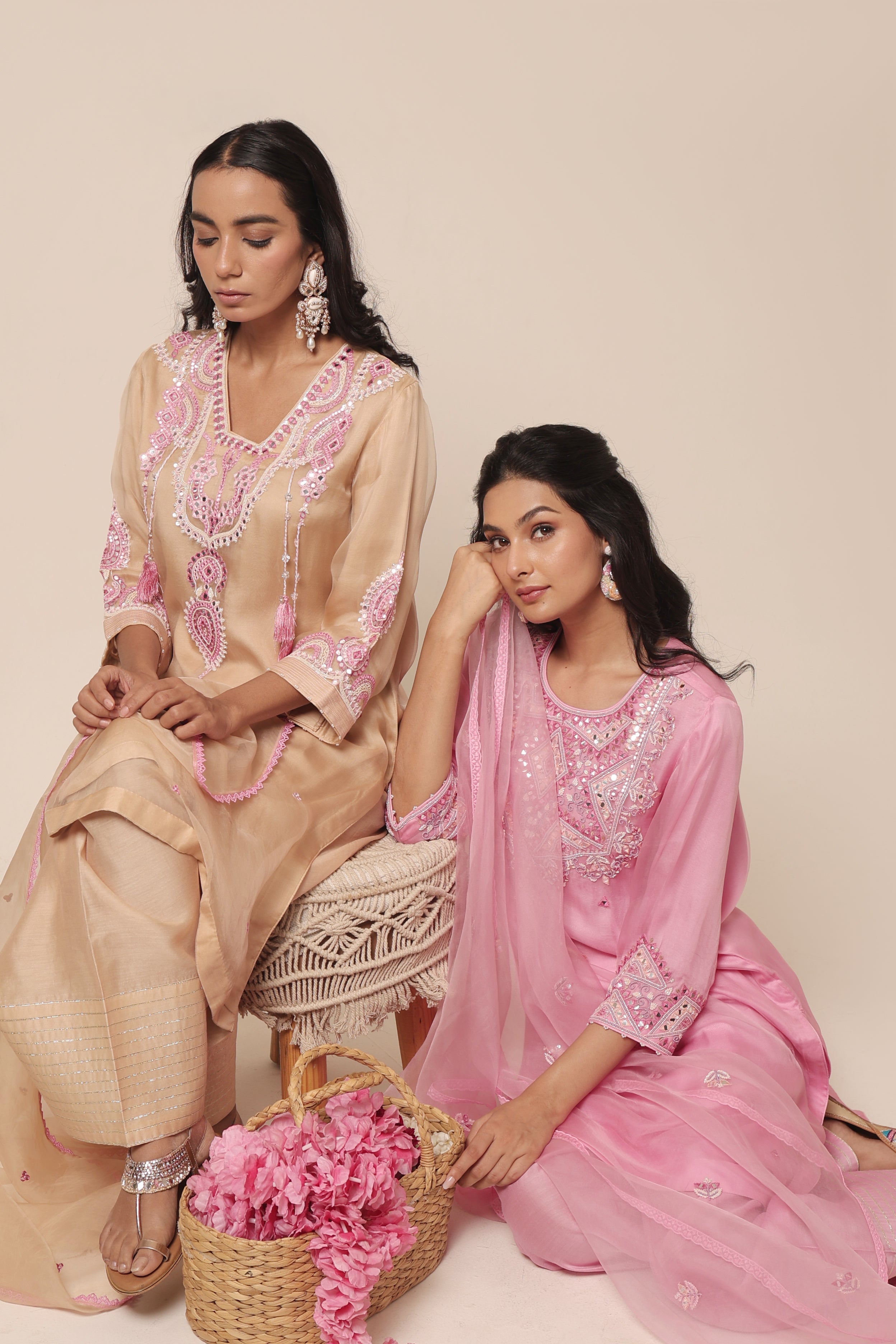 Two women in traditional outfits with intricate embroidery, one in beige and the other in pink, sitting on a white surface with a beige background.