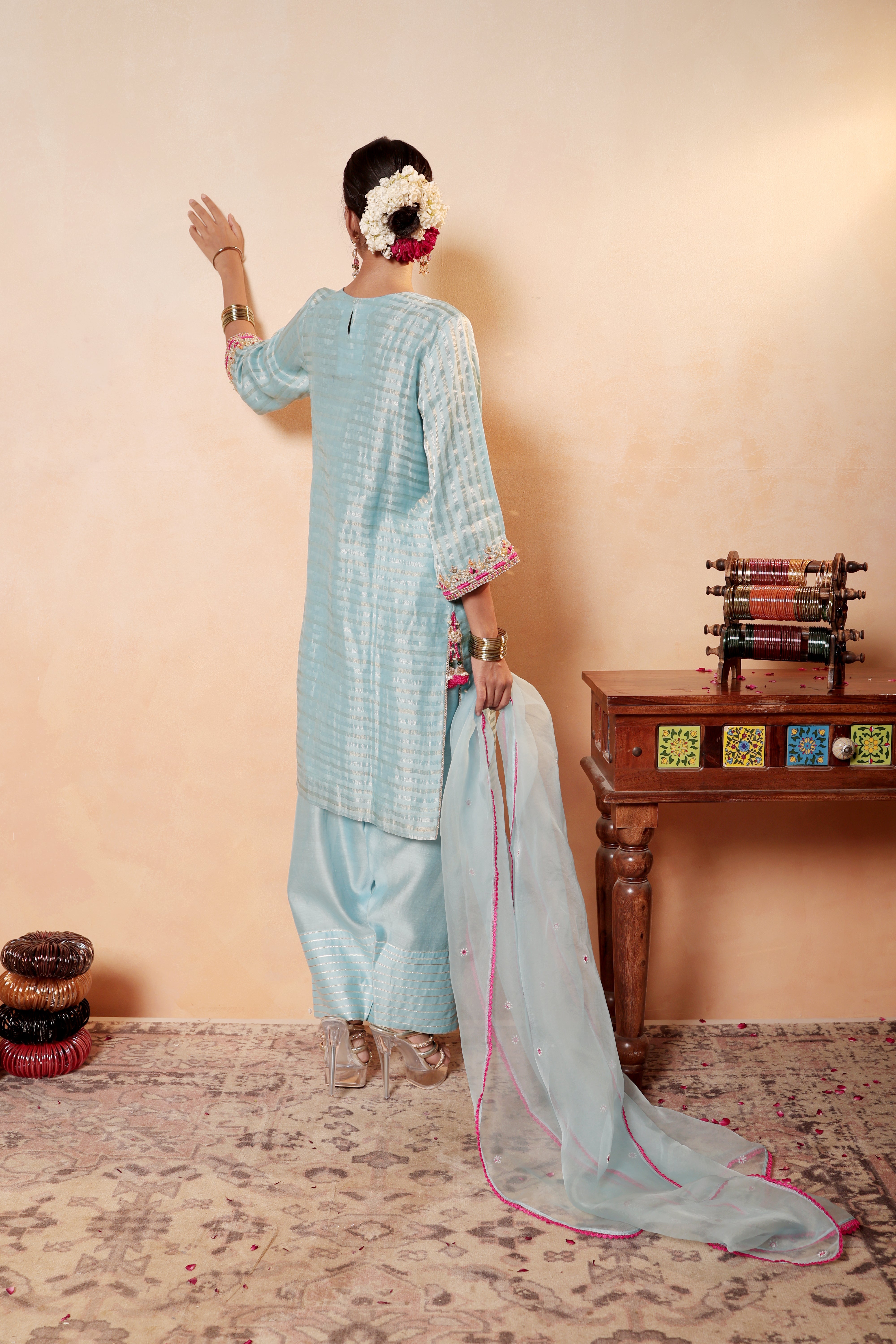 Woman in a light blue traditional outfit with a pink border, standing against a beige wall. Model wearing sky blue silk striper tissue kurta with gota embroidery, styled with traditional farshi salwar and dupatta – Sunehri Gota Collection by House of AAS.
